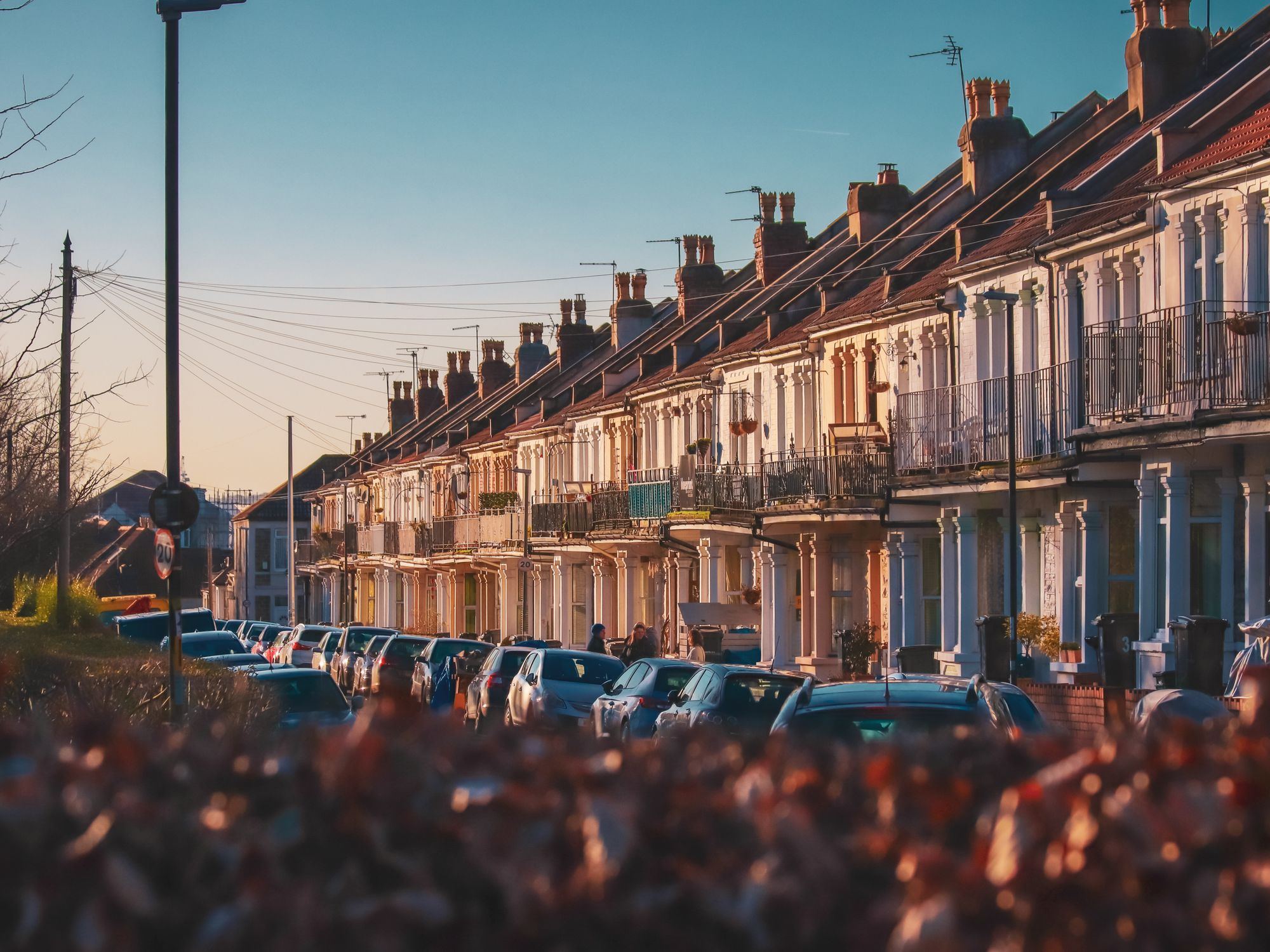 A row of Victorian terraced brick houses with cars parked along the street at sunset.