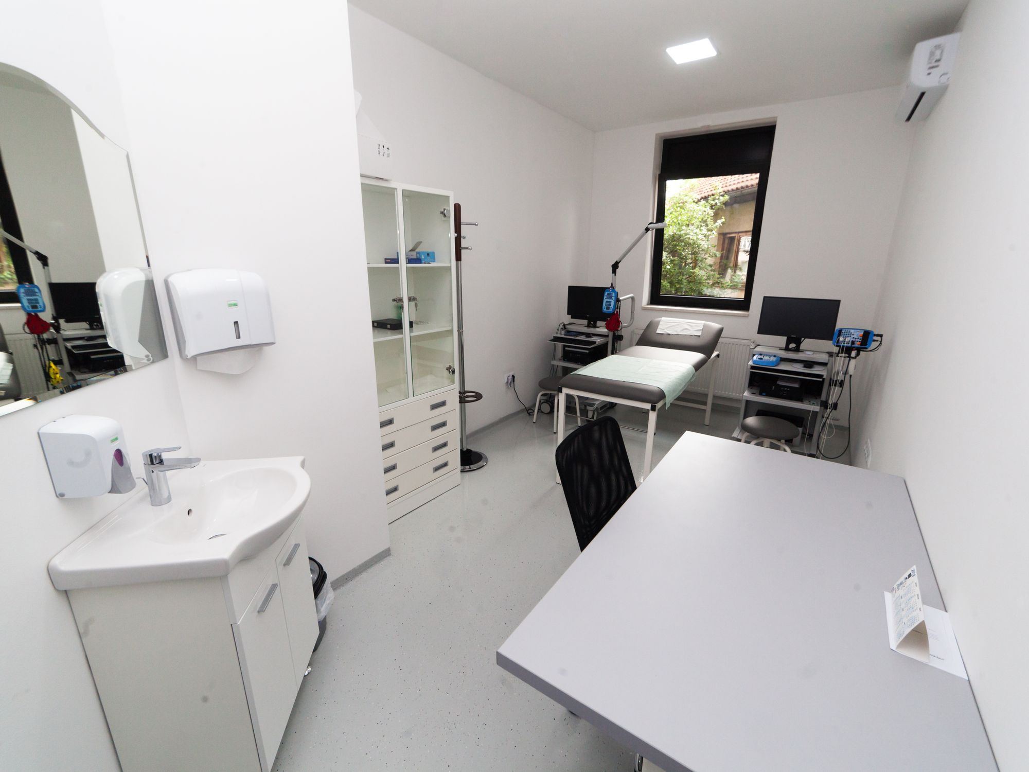 A bright clean medical examination room with a bed, desk and storage cabinet.